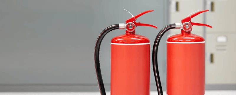 Two red fire extinguishers with hoses standing side by side on a white surface in a maintenance area.