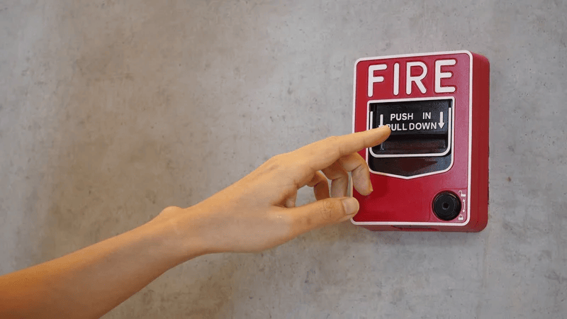Person presses a red wall-mounted fire alarm pull station labeled FIRE, pushing in and pulling down to activate.