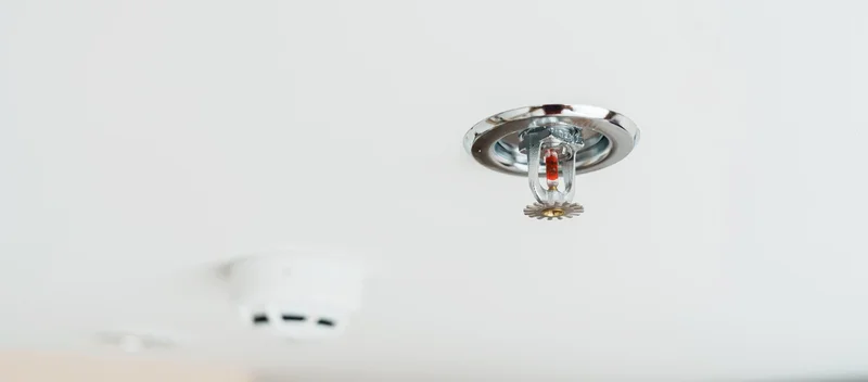 Bare ceiling light fixture with exposed bulb hanging from a recessed socket; white ceiling and a distant smoke detector in the background.
